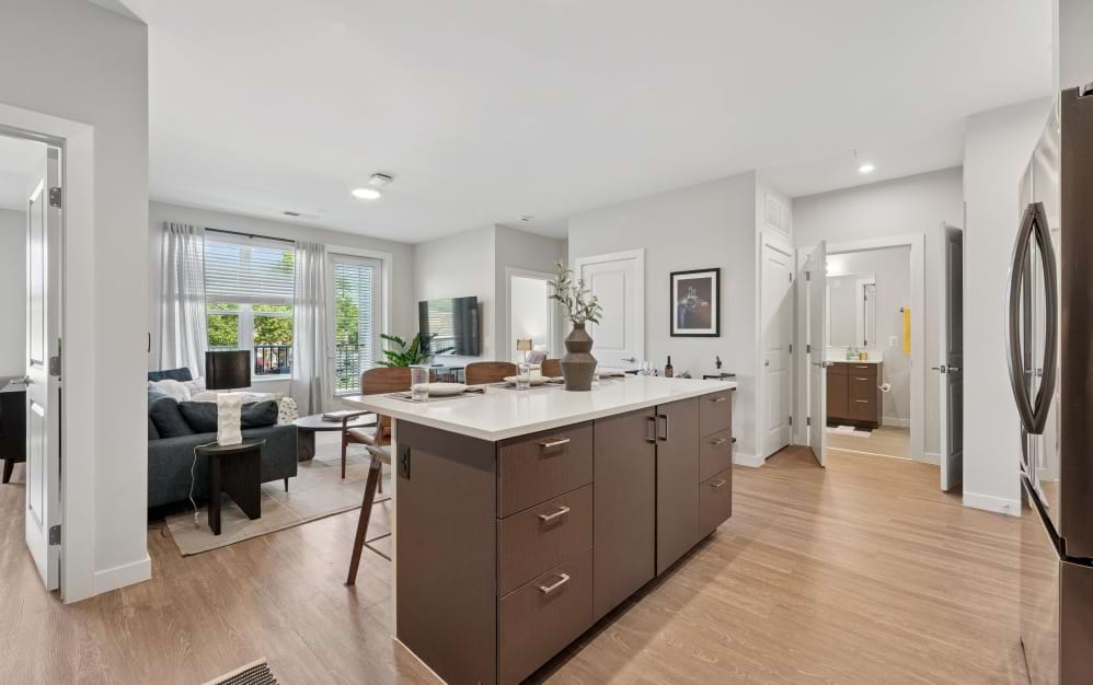 Contemporary A9 kitchen at Alta NOVA featuring a large white countertop island, wood cabinetry, wood-style flooring, and an adjoining living room with natural light.