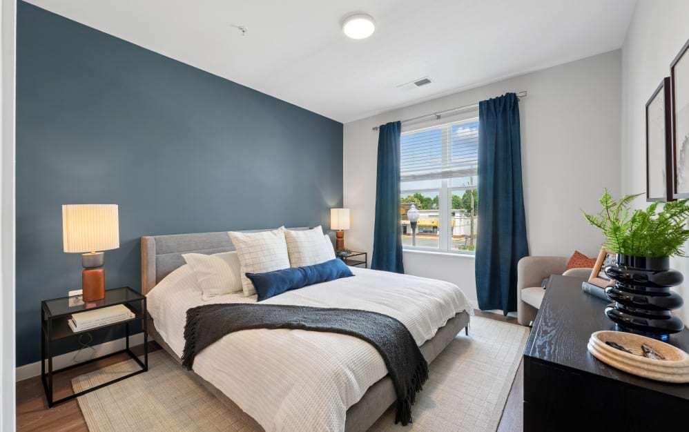 Spacious A9 bedroom at Alta NOVA apartments in Falls Church, VA, featuring a blue accent wall, upholstered bed, modern furnishings, wood-style flooring, and a large window with natural light.