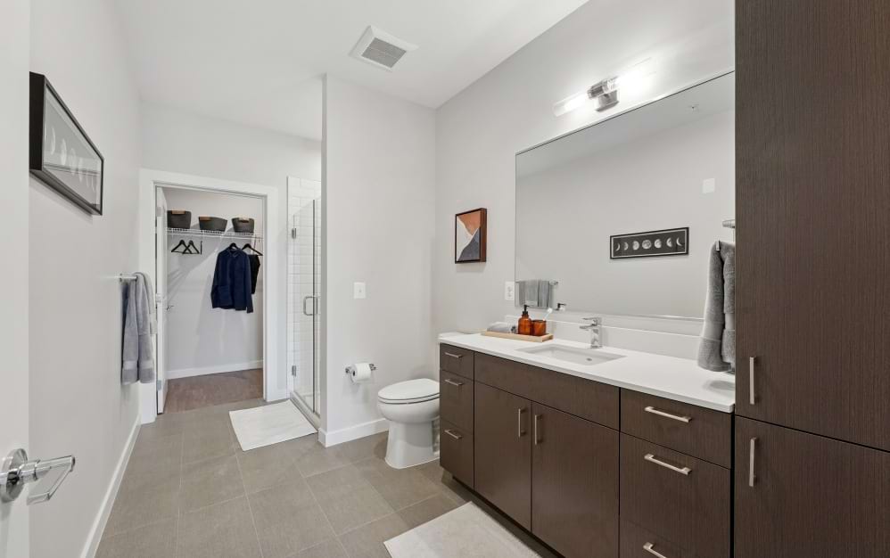 Modern A9 apartment bathroom at Alta NOVA featuring warm wood cabinetry, white countertops, a spacious vanity, and a glass-enclosed shower with white subway tile.