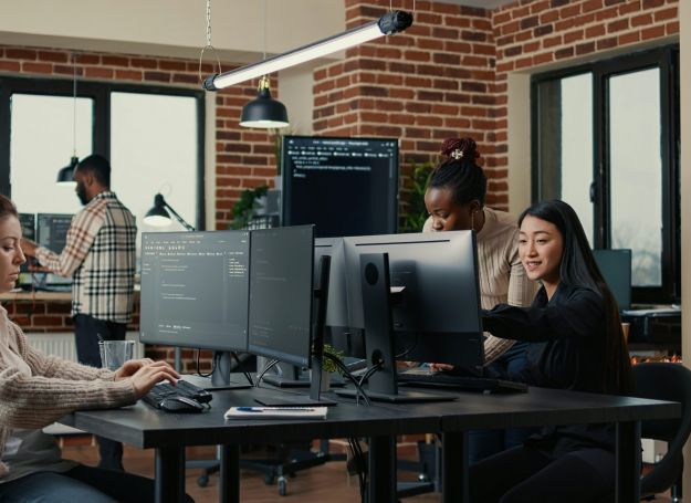 Four people work on computers in a modern office with brick walls, reminiscent of the stylish interiors at Alta Nova apartments. One person types while another points at a screen.