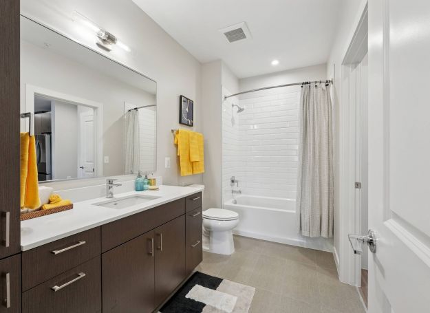 Alta Nova apartments Modern bathroom with double sink, dark cabinets, yellow towels, bathtub with white curtain, and large mirror.