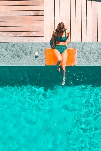 A woman sits at the pool's edge in a green bikini, her feet dipping into the water. Wooden decking frames the serene blue pool below, creating a picturesque setting at Alta Nova apartments.