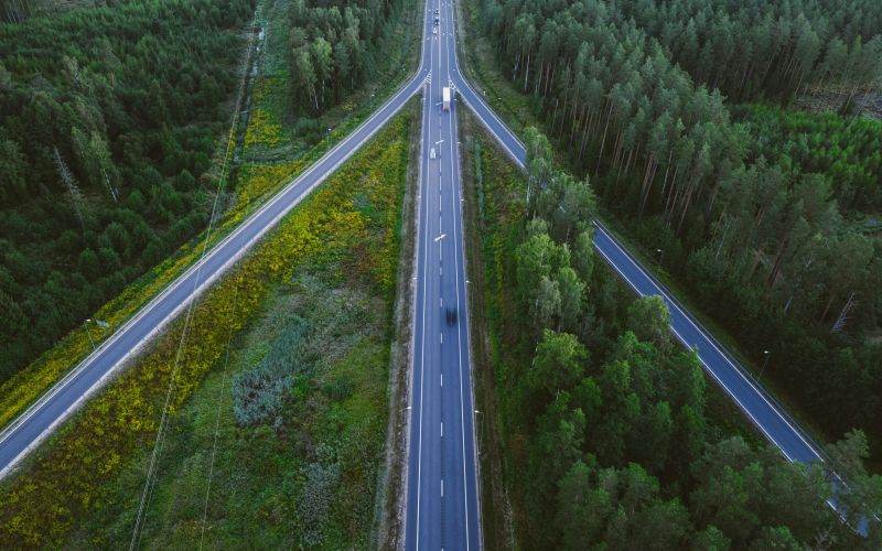 Aerial view of a T-shaped intersection surrounded by dense green forest and some yellow wildflowers, with the Alta Nova apartments subtly peeking through the foliage.