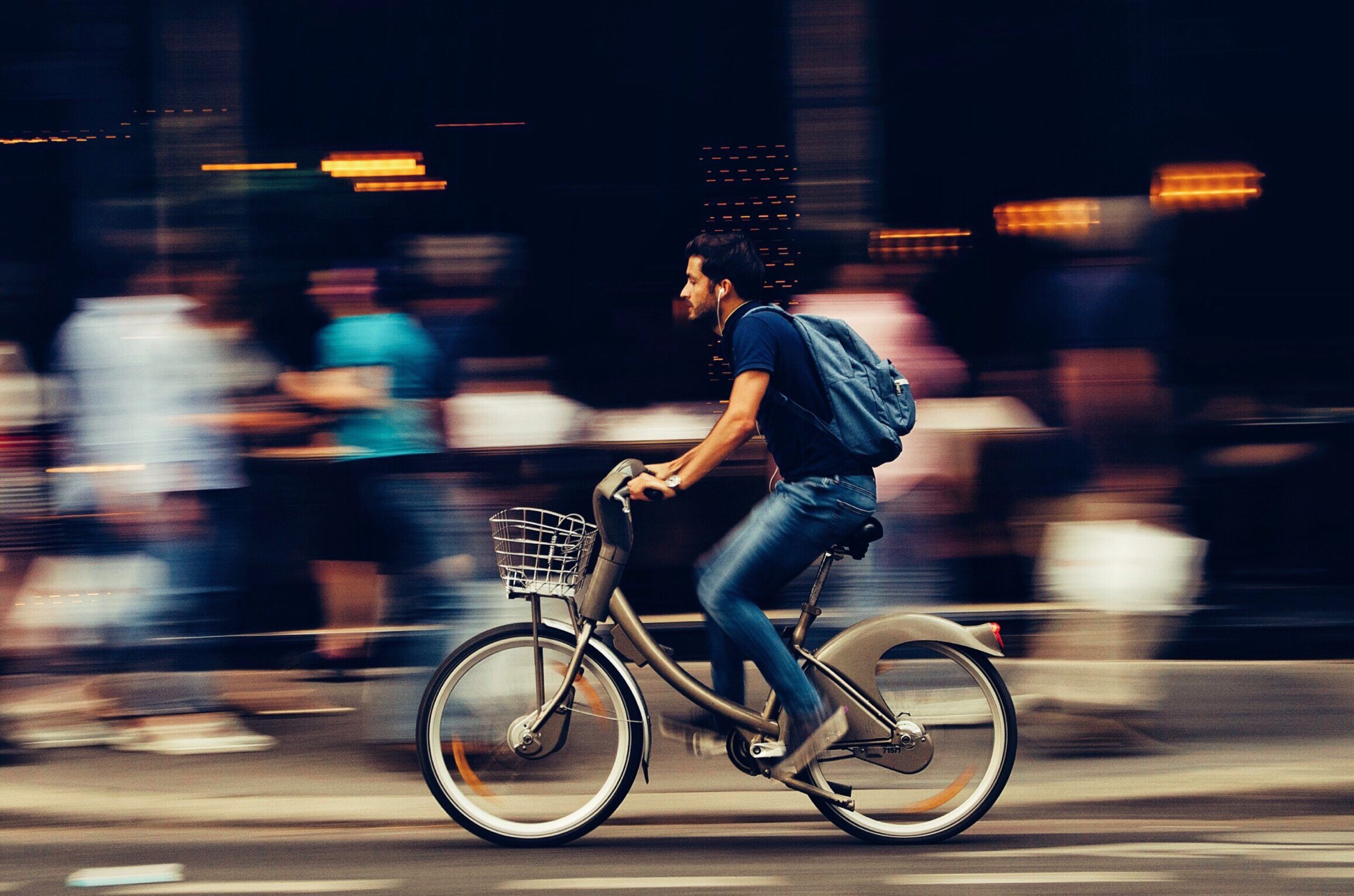 A man riding a bicycle quickly through a busy urban street with the blurred pedestrian background and the towering Alta Nova apartments looming in the distance.