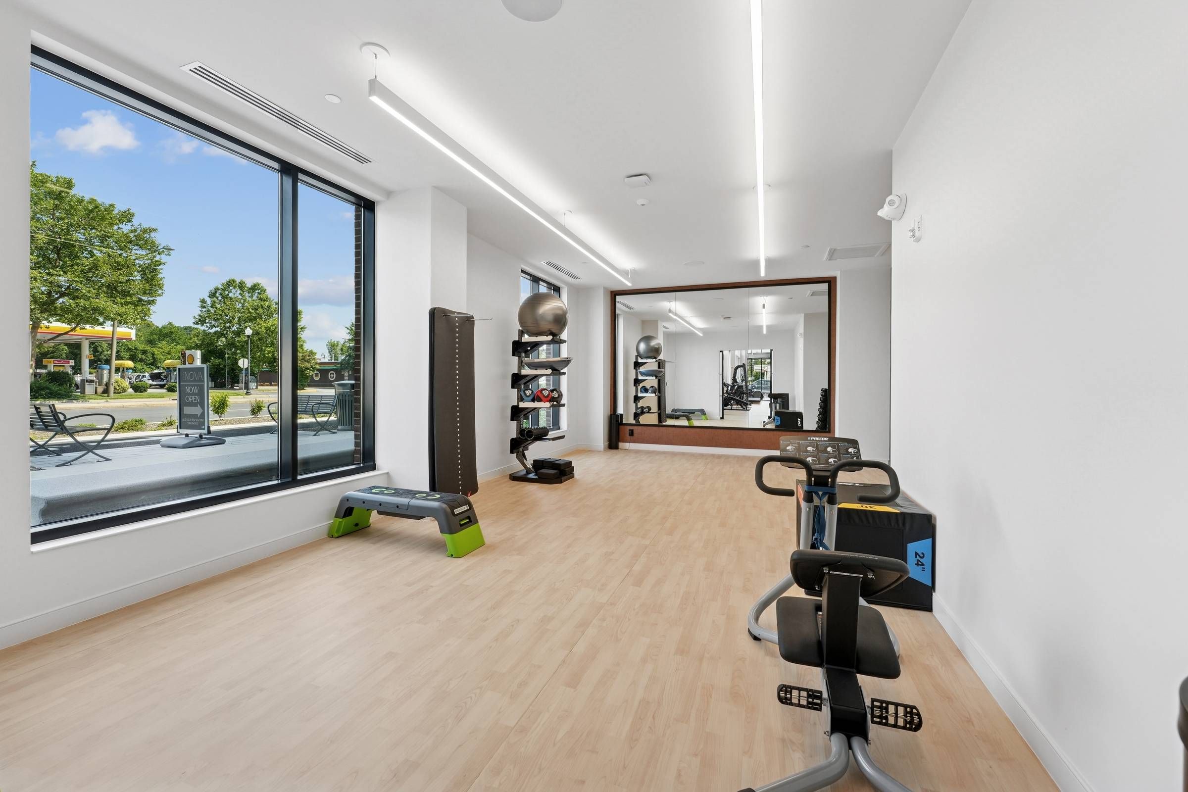 Alta Nova apartments Modern gym room with exercise equipment, large mirror, and big window showing trees and outdoor seating.