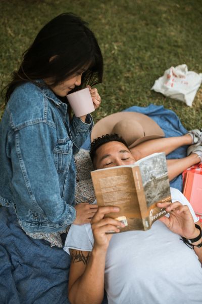 A person leans over with a cup while another lies on a blanket reading a book outdoors, enjoying the serene ambiance of Alta Nova apartments.