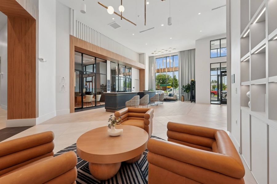 Alta Nova apartments Modern lobby with tan leather chairs, round table, large windows, and a view of a seating area and greenery outside.